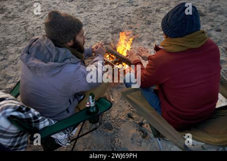 Campfire, friends and talking with men by the beach at sunset with vacation and camping. Ocean, outdoor and together with travel and people back on Stock Photo
