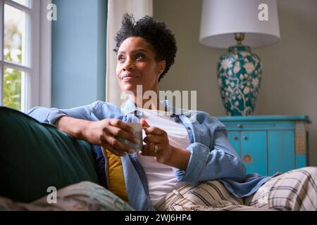 Mature Woman At Home Drinking Coffee Relaxing On Sofa In Lounge Stock Photo