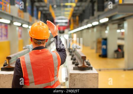 Rear view of electric train industry engineer wearing a safety helmet and reflective vest. There is an electric car in the car r Stock Photo
