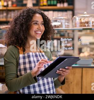 Smiling Young Woman Wearing Apron Working In Food Shop Using Digital Tablet Stock Photo
