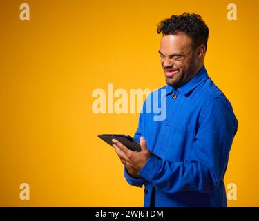 Studio Portrait Of Man With Down Syndrome Using Digital Tablet Against Yellow Background Stock Photo