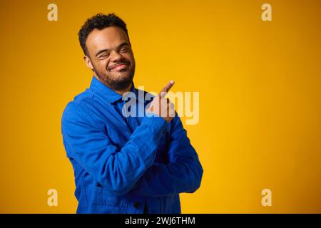 Studio Portrait Of Smiling Man With Down Syndrome Standing Against Yellow Background Pointing Stock Photo