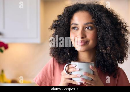 Smiling Young Woman In Kitchen At Home Taking A Break Holding Hot Drink Stock Photo