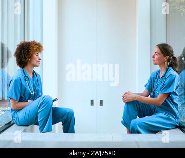 Two Female Doctors Wearing Scrubs Taking A Break In Modern Hospital Stock Photo