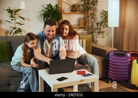 Young family, father, other and little daughter sitting at home with packed suitcases and booking trip online via laptop Stock Photo