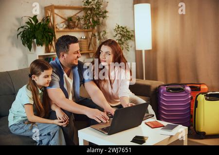 Young family, father, other and little daughter sitting at home with packed suitcases and booking trip online via laptop Stock Photo