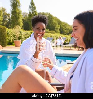 Two Mature Female Friends Wearing Robes Outdoors On Loungers Drinking Champagne On Spa Day Stock Photo
