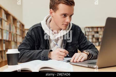 Male student with ginger hair preparing for an exam while sitting in a library. High school student sitting at a table with laptop. Stock Photo