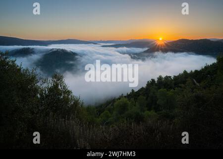 The sun rises in a clear sky over the Judea mountains in Israel, as clouds fill the valleys among them. Jerusalem may be seen in the hazy horizon. Stock Photo