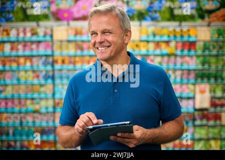 Mature Man Working Indoors In Garden Centre Using Digital Tablet In Front Of Display Of Seeds Stock Photo
