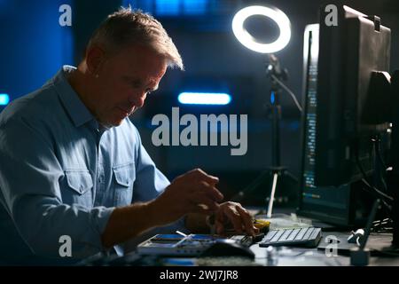 Mature Male Electronics Expert Repairing Laptop In Workshop With Low Key Lighting Stock Photo