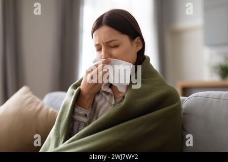 Sick young woman wrapped in blanket blowing her nose Stock Photo