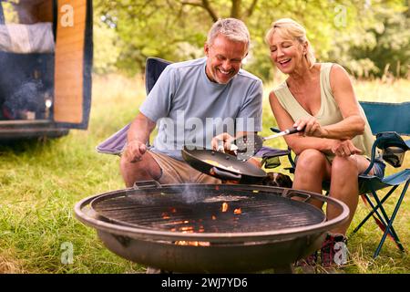 Senior Couple Camping In Countryside With RV Cooking Bacon And Eggs For Breakfast Outdoors On Fire Stock Photo