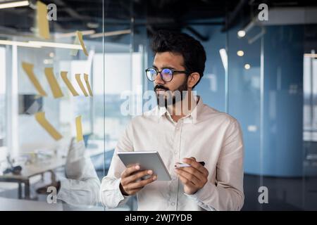 Serious and focused young Indian man working in office with tablet, standing near glass board with notes, processing data, thinking about project. Stock Photo