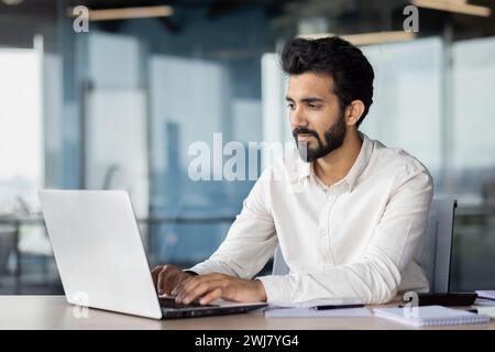 A serious and focused young Indian man is working on a laptop in the office. He sits at his desk and types on the keyboard. Stock Photo