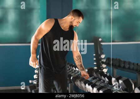 Athletic young man carefully selects dumbbells from the rack at gym Stock Photo