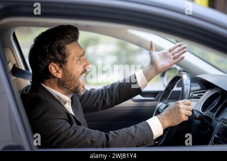 Close-up photo of angry young businessman in suit driving car and emotionally gesturing with hands, displeased shouting and arguing. Stock Photo
