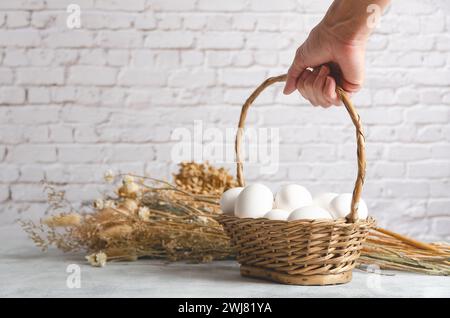 Female hand holding a basket with white chicken eggs on a light background with dried flowers. Front view with copy space. Stock Photo