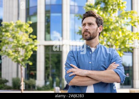 Mature bearded man in casual shirt standing with folded arms, looking thoughtful outside an office building. Stock Photo