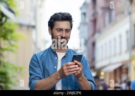Bearded mature male tourist in casual attire using his smartphone while walking on urban street during evening time. Stock Photo