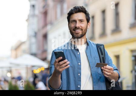 Portrait of a young male tourist standing on a city street with a backpack, holding a credit card and a mobile phone. Smiling and looking at the camera. Stock Photo