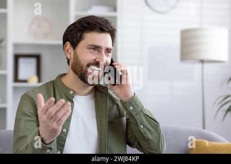 Cheerful bearded middle-aged man enjoying a phone call while sitting on a sofa in a bright, modern living room setting. Stock Photo