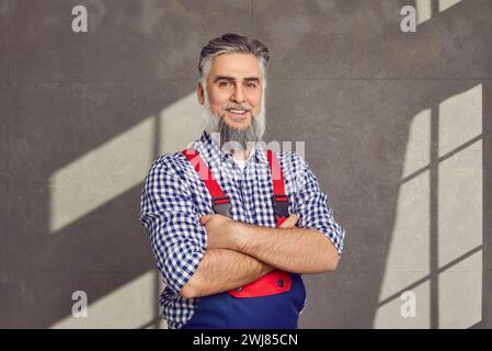 Portrait of a happy smiling mature bearded repairman on grey background in overall. Stock Photo