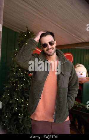 Portrait of happy handsome man in trendy jacket and sunglasses standing near Christmas tree. Fashionable male hipster posing with hand in hair and smiling. Stock Photo