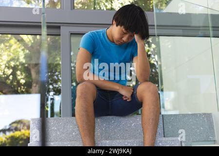 Teenage Asian boy sits outside a modern building, looking thoughtful and sad Stock Photo