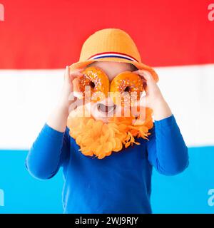 Little Dutch boy girl wearing country symbols eating orange donuts celebrating King day. Children support Holland sport team. Kids from the Netherland Stock Photo