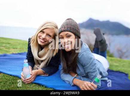 Picnic, grass and portrait of women in field with smile, happy and relaxing on weekend outdoors. Friends, countryside and people with drinks for Stock Photo
