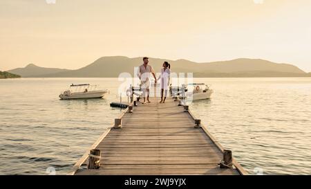 couple walking on a wooden pier in the ocean at sunset in Thailand.Caucasian men and Asian woman diverse couple waking at a jetty in the ocean, man and women watching sunset together Stock Photo