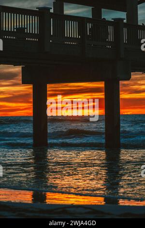 A breathtaking view of a vibrant sunset with fiery hues reflecting on the water under a pier on a beach in Miami, Florida. Stock Photo