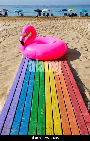 Inflatable pink flamingo tube on wooden floor of purple blue green yellow orange and red on sandy beach against people sitting under outdoor umbrellas Stock Photo