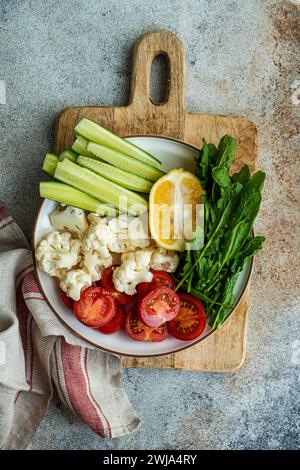 Top view of vibrant dish of raw vegetables with sliced cucumber, halved cherry tomatoes, cauliflower pieces, a lemon slice, and arugula on a cutting b Stock Photo