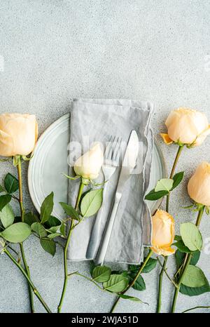 From above sophisticated table arrangement featuring a plate with a linen napkin, silverware, and peach roses spread around on concrete background Stock Photo