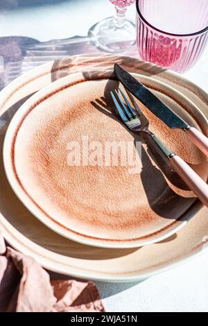 An elegant table setting featuring textured ceramic plates, modern cutlery, and pink glassware, casting soft shadows in sunlight. Stock Photo