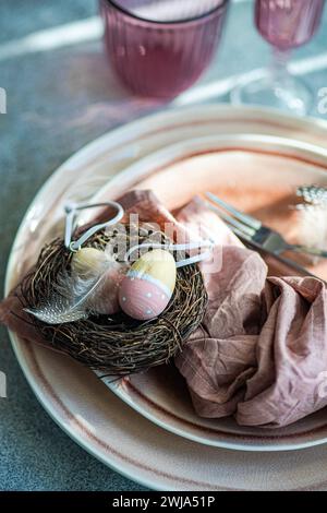 An artistic Easter table setting featuring a nest with pastel decorated eggs atop ceramic plates, complemented by a dusty pink napkin. Stock Photo