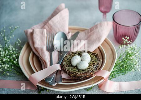 Elegant Easter table setting with pastel-colored napkins, plates, and decorative eggs nested in a twig nest, complemented by flowers and ribbon. Stock Photo