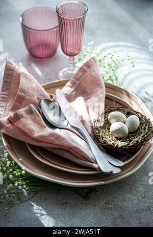 An elegant Easter dining setup featuring a nest with speckled eggs, pink napkins, and matching colored glassware on a textured surface. Stock Photo