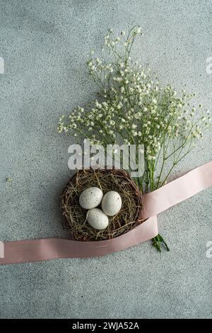 Top view of speckled Easter eggs nestled in a rustic basket, adorned with soft-pink ribbon and delicate white flowers, arranged on a speckled gray bac Stock Photo