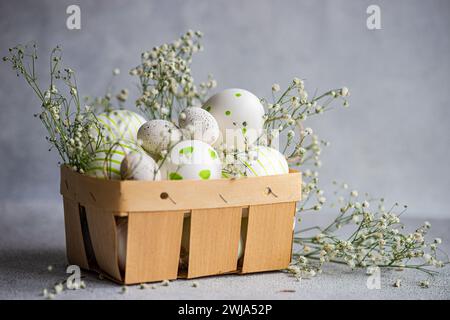 A wooden basket filled with patterned Easter eggs, adorned with delicate gypsophila flowers on a textured background Stock Photo