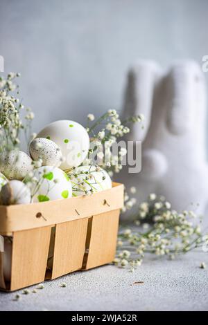 A selection of decorated Easter eggs nestled in a wooden basket alongside sprigs of gypsophila, with a soft gray background Stock Photo