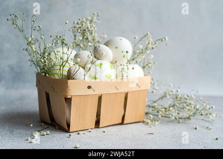 A wooden basket filled with decorated Easter eggs among delicate white spring flowers, on a neutral background Stock Photo