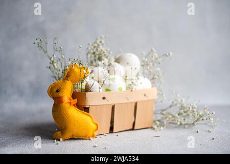 A plush yellow Easter bunny gazes at a basket filled with decorated white eggs and sprigs of baby's breath on a textured grey background Stock Photo
