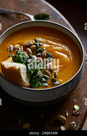 A warm bowl of creamy pumpkin soup garnished with pumpkin seeds, pistachios, and vibrant green pesto, served on a rustic wooden surface Stock Photo