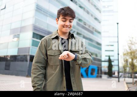 Self assured male in t shirt and jacket with toothy smile standing and looking at smart watch against modern building on street in daytime Stock Photo