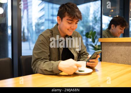 Positive young brunet male in stylish outfit sitting in cafe using smartphone and drinking coffee while looking at screen Stock Photo