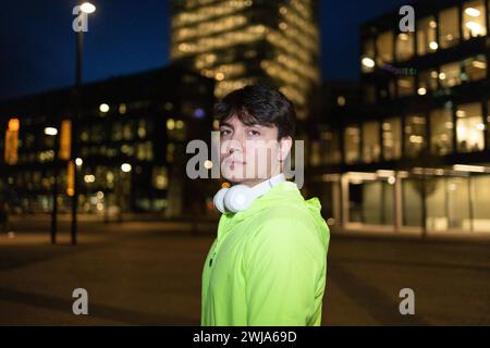 Side view of confident young male in casual jacket and headphones around neck standing on city street at night while looking at camera Stock Photo