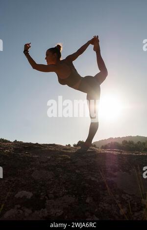Full body of flexible barefooted female in activewear practicing Lord of the Dance yoga pose during fitness training on rocky cliff against sunset sky Stock Photo
